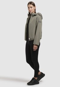 Lightweight, olive green jacket with a hood, zip closure, and front logo. Worn over a black top and leggings, paired with black sneakers.