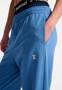Lysblå joggers med elastisk talje, sidelommer og et lille hvidt chevronlogo på venstre ben. Blød stofstruktur.