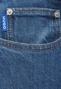 Τσέπη τζιν σε μπλε denim με χρυσαφένιες ραφές, διακοσμημένη με μπλε ετικέτα "HUGO" και ασημένιο κουμπί, που παρουσιάζει υφασμάτινη υφή.