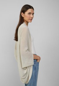 Leichter, offen gestrickter Cardigan in Creme, mit weiten Ärmeln, lockerer Drapierung und strukturiertem Finish. Kombiniert mit einem weißen Oberteil und Jeans.