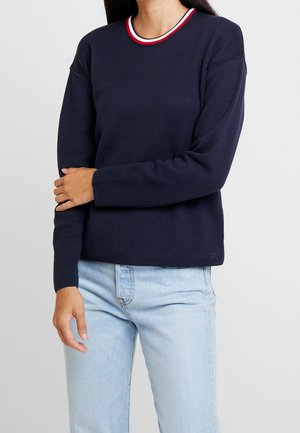 Frau trägt einen navyblauen Pullover mit einem rot-weiß-blau gestreiften Kragen und hellblauer Jeans, steht mit einem Arm verschränkt.