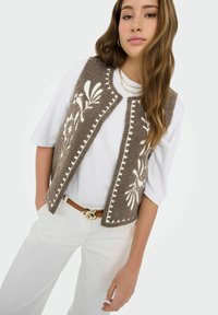 Jeune femme portant une chemise et un pantalon blancs avec un gilet marron texturé orné de broderies florales blanches et une ceinture marron à boucle dorée.