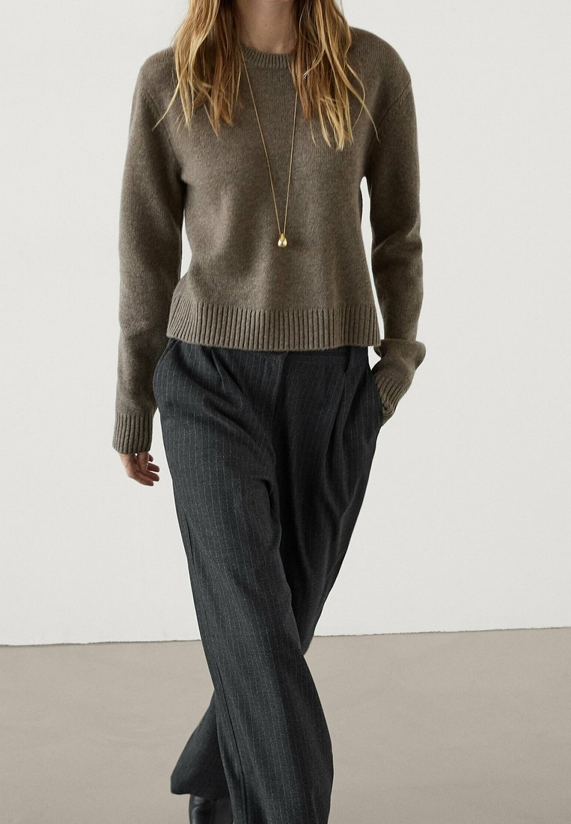 Maglione di lana marrone con orlo a coste, abbinato a pantaloni scuri a righe sottili. Maniche lunghe evidenti e collana con ciondolo dorato.