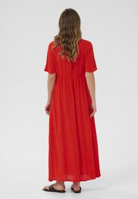 Robe maxi rouge à manches courtes, taille cintrée et jupe fluide. Tissu léger avec une surface légèrement texturée. Sandales noires.