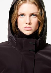 Giacca impermeabile viola scuro con colletto alto, cappuccio attaccato e dettagli neri. Texture liscia e design minimalista.