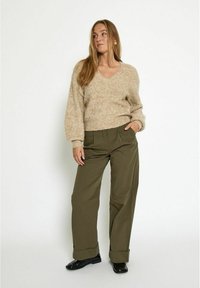 Beige, fuzzy gebreide trui met een V-hals; loszittende olijfgroene broek met plooien en omgeslagen uiteinden; zwart glanzende schoenen.