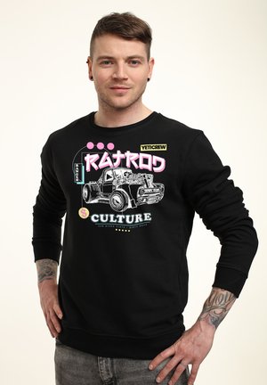 Young man with short hair poses wearing a black sweatshirt featuring a vintage truck illustration and colorful stylized text.