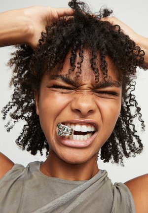 Cabello rizado y negro, camiseta sin mangas gris, sonriendo con un anillo de plata que tiene piedras preciosas verdes en la boca, expresión facial que muestra esfuerzo.