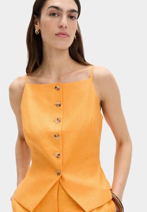 Frau trägt ärmelloses oranges Top mit sechs Knöpfen vorne und dünnen Schulterriemen, Hände in den Taschen, neutraler Hintergrund.