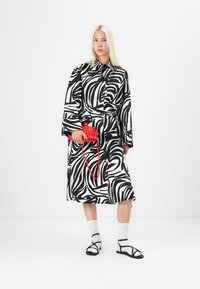 Rochie cămașă cu model de zebră alb-negru, cu un cordon, asortată cu sandale negre și șosete albe; ținând o geantă roșie.