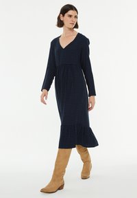 Largo vestido azul marino con escote en V, mangas largas y dobladillo acampanado. Presenta un patrón de cuadros y una tela suave y texturizada. Se lleva con botas de rodilla color marrón claro.