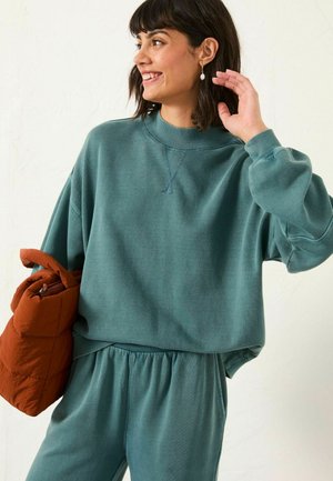 Türkiser Baumwoll-Sweatshirt mit geripptem Rundhalsausschnitt und Bündchen, kombiniert mit passenden Jogginghosen. Das Modell hält eine gesteppten orangen Tasche.