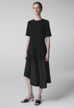 Junge Frau steht, trägt ein schwarzes Midi-Kleid mit kurzen Ärmeln und schwarze offene, hochhackige Mules mit goldenem Kugeldetail an den Absätzen.