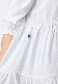 Weißes Kleid mit dezenten vertikalen Streifen, gerafften Ärmeln und einem gestuften Saum. Ein kleines blauer Tag mit der Aufschrift 'SEASIDE' ist an der Seitennaht sichtbar.