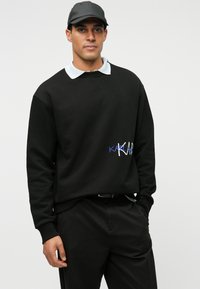 Man i svart keps, svart sweatshirt med vit och blå text på bröstet, vit skjorta under, och svarta byxor, står mot en vit bakgrund.