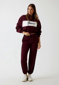 Bordeauxrotes Sportset mit einem gekürzten Sweatshirt mit weißem Band und gesticktem "Guess"-Logo, kombiniert mit passenden, schmal zulaufenden Hosen.