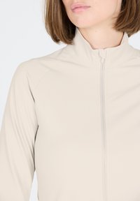 Beige Zip-Jacke mit hohem Kragen, langen Ärmeln und glatter Textur sowie subtilen Nähdetails entlang des Reißverschlusses.