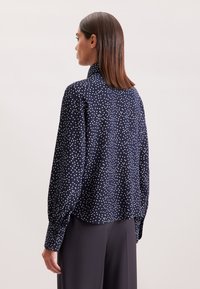 Blusa a pois blu navy con colletto alto, maniche lunghe e polsini abbottonati. Tessuto morbido con vestibilità ampia e orlo arrotondato.