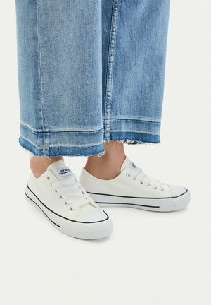 Białe niskie trampki z płótna z czarną lamówką noszone z niebieskimi jeansami o skróconych, postrzępionych nogawkach na jednolitym białym tle.