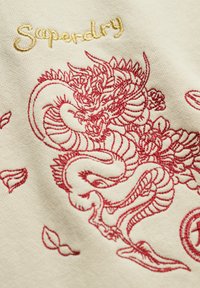 Rode geborduurde draak met bloemrijke details en gouden "Superdry" tekst op beige stof.