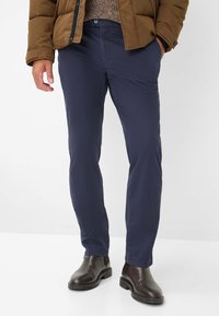 Marineblaue, eng geschnittene Baumwollhosen mit glatter Textur, Eingrifftaschen und Knopfverschluss, kombiniert mit braunen Lederstiefeletten.