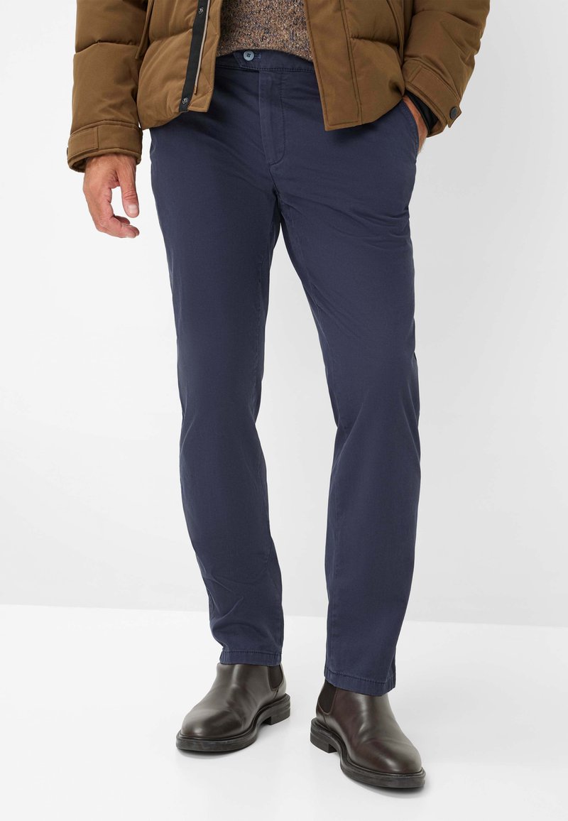 Marineblaue, eng geschnittene Baumwollhosen mit glatter Textur, Eingrifftaschen und Knopfverschluss, kombiniert mit braunen Lederstiefeletten.