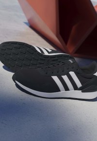 Sneakers Adidas nere con strisce bianche poggiate su una superficie in cemento accanto a una struttura geometrica rossa.