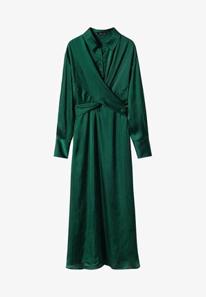 Kvinde iført en langærmet, dybgrøn midi slå-om kjole kombineret med sorte, snørede hæle og stående på et neutralt gulv.