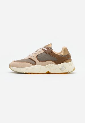 Vista lateral de una zapatilla deportiva beige y marrón con suela blanca y cierre con cordones sobre fondo blanco.