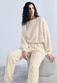 Γυναίκα με σγουρά σκουρόχρωμα μαλλιά καθισμένη σε στρογγυλό σκαμπό με υφή, φοράει κρεμ φλις πουλόβερ με πλεκτό σχέδιο κοτσίδας και ταιριαστό παντελόνι.