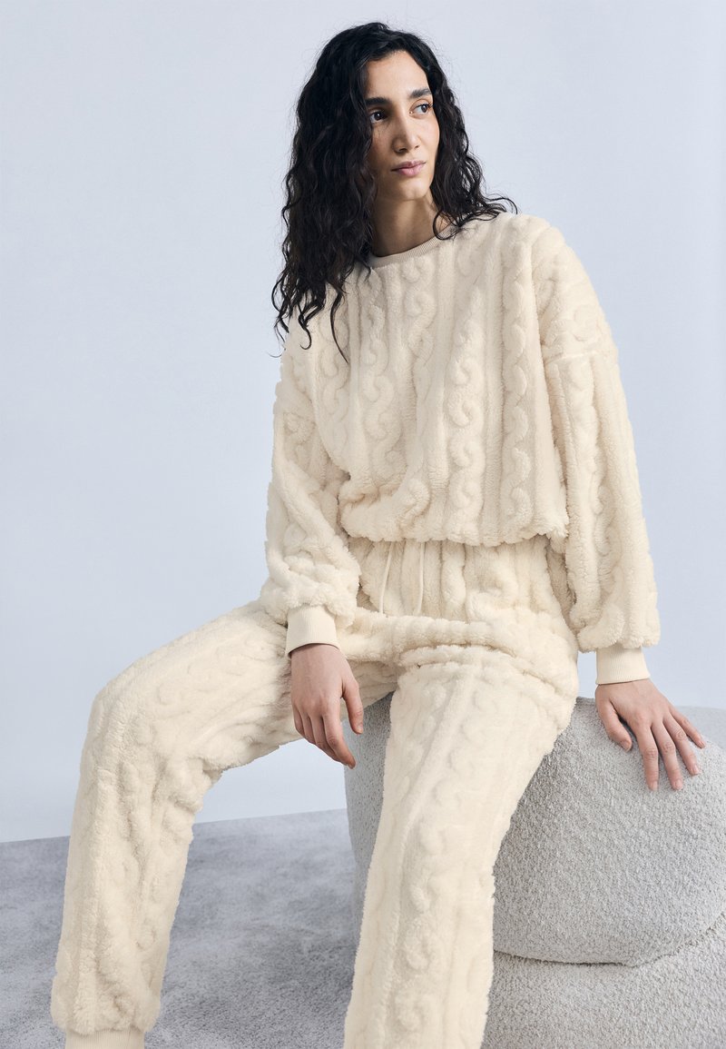 Γυναίκα με σγουρά σκουρόχρωμα μαλλιά καθισμένη σε στρογγυλό σκαμπό με υφή, φοράει κρεμ φλις πουλόβερ με πλεκτό σχέδιο κοτσίδας και ταιριαστό παντελόνι.