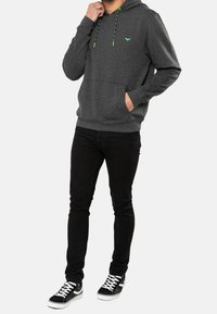 Personne portant un sweat à capuche gris foncé avec des cordons verts et un emblème, un jean noir slim et des baskets noires à semelles blanches et lacets blancs.