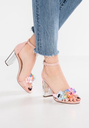 Voeten in roze sandalen met hoge hakken, enkellinten en meerkleurige bloemversieringen, gecombineerd met blauwe jeans met versleten zoom.