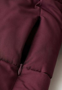 Chaqueta de plumas burdeos con una textura suave, que presenta un detalle de bolsillo con cremallera en el lado izquierdo.