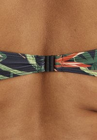 Close-up af en persons ryg, der bærer en mørk bikini top med tropisk mønster og en sort plastiklukning i midten.