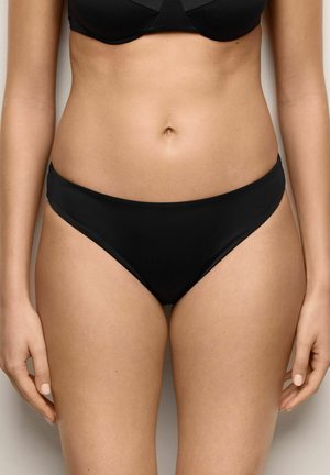 Kobieta w czarnym bikini na dole i górze, stojąca na neutralnym tle z ramionami swobodnie opuszczonymi wzdłuż ciała.