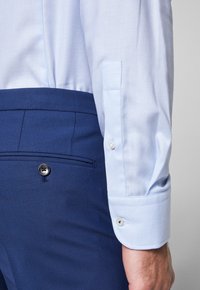Camicia azzurra chiara con texture e polsini abbottonati, abbinata a pantaloni su misura blu navy con dettaglio a bottoni e frontale piatto.