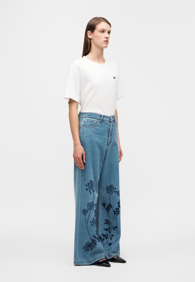 Λευκό υπερμεγέθες t-shirt με μικρό λογότυπο, συνδυασμένο με φαρδιά μπλε τζιν παντελόνια που διαθέτουν μαύρη κεντητή λουλουδένια διακόσμηση στο κάτω μέρος της πλευράς. Μαύρα μυτερά παπούτσια.