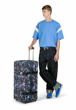 Bolsa de lona negra con ruedas y patrón abstracto colorido, tela duradera, asas dobles y correas, junto a una persona con camisa azul y vaqueros oscuros.