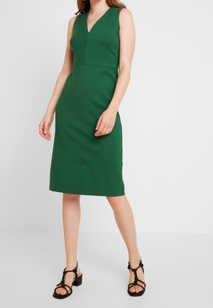 Femme portant une robe verte sans manches au genou avec un décolleté en V et des sandales noires à talons blocs à brides, debout sur fond blanc.