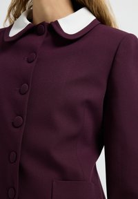 Manteau bordeaux avec un col Peter Pan blanc, doté de boutons ronds, d'une texture lisse et d'un détail de poche à l'avant.