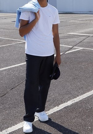 Personne debout dans un parking vide, portant un t-shirt blanc, un pantalon noir, des baskets blanches, tenant une casquette dans une main et une veste bleue sur l'épaule.