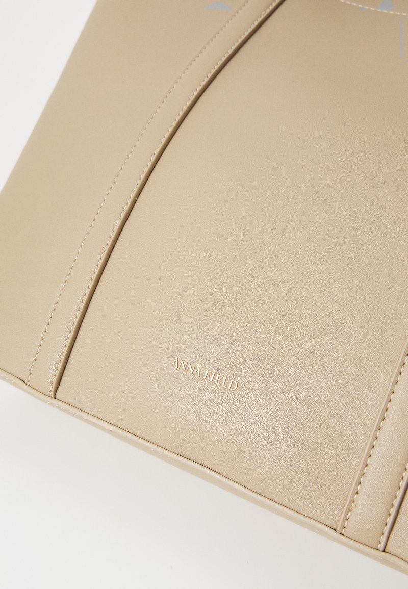 Bolsa tote de cuero sintético beige con acentos cosidos y un logotipo en relieve dorado, con una textura suave y un diseño estructurado.