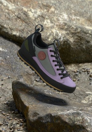 Chaussure de randonnée violet clair et gris avec semelle et lacets noirs flottant au-dessus d’un terrain rocheux, avec un patch rond marron du logo sur le côté.