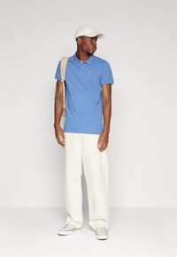 Camisa polo azul de manga corta y cuello, pantalones blancos, zapatillas grises y un bolso beige. Una gorra blanca completa el atuendo.