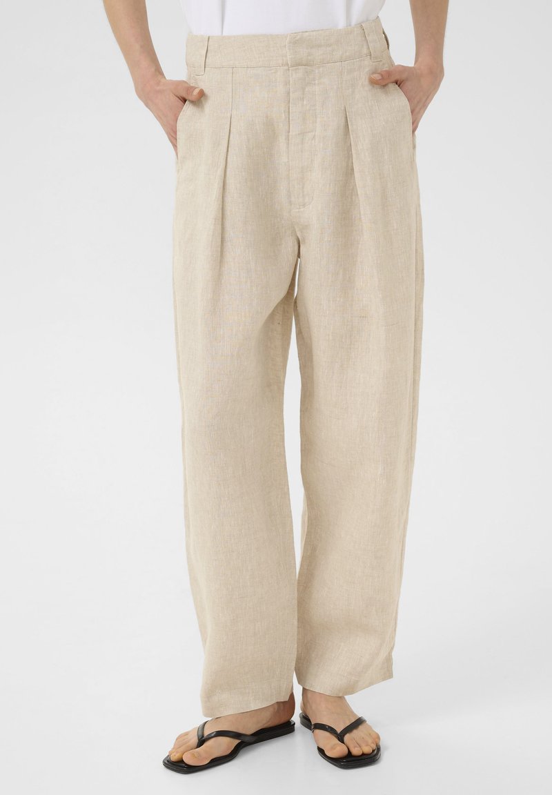 Beige linnen broek met een wijd ontwerp, voorzien van een geplooide voorkant en zijzakken. Gemaakt in combinatie met zwarte teenslippers, wat zorgt voor een ontspannen pasvorm.