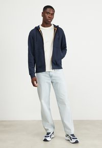 Marineblauwe zip-up hoodie met contrasterende oranje voering, gecombineerd met lichtblauwe jeans en witte sneakers. Glad materiaal en een losse pasvorm.