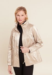 Giacca metallica oro con colletto alto, due tasche frontali e chiusura con zip. Il tessuto ha una finitura lucida e texture. Top e pantaloni neri.