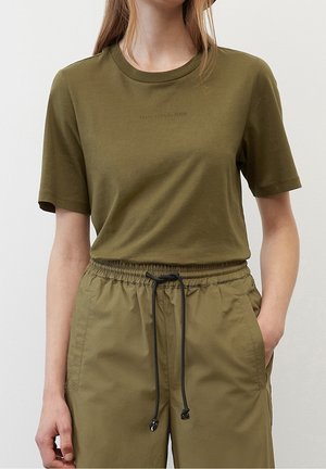 Kvinde iført olivengrøn T-shirt med korte ærmer og matchende bukser med elastik i taljen og sort snor, stående med den ene hånd i lommen.