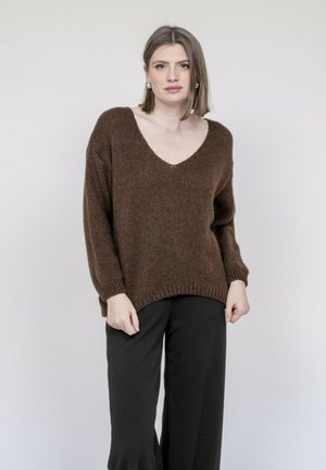 Femme aux cheveux bruns clairs et raides portant un pull marron à col en V oversize et un pantalon large noir, debout devant un fond clair uni.